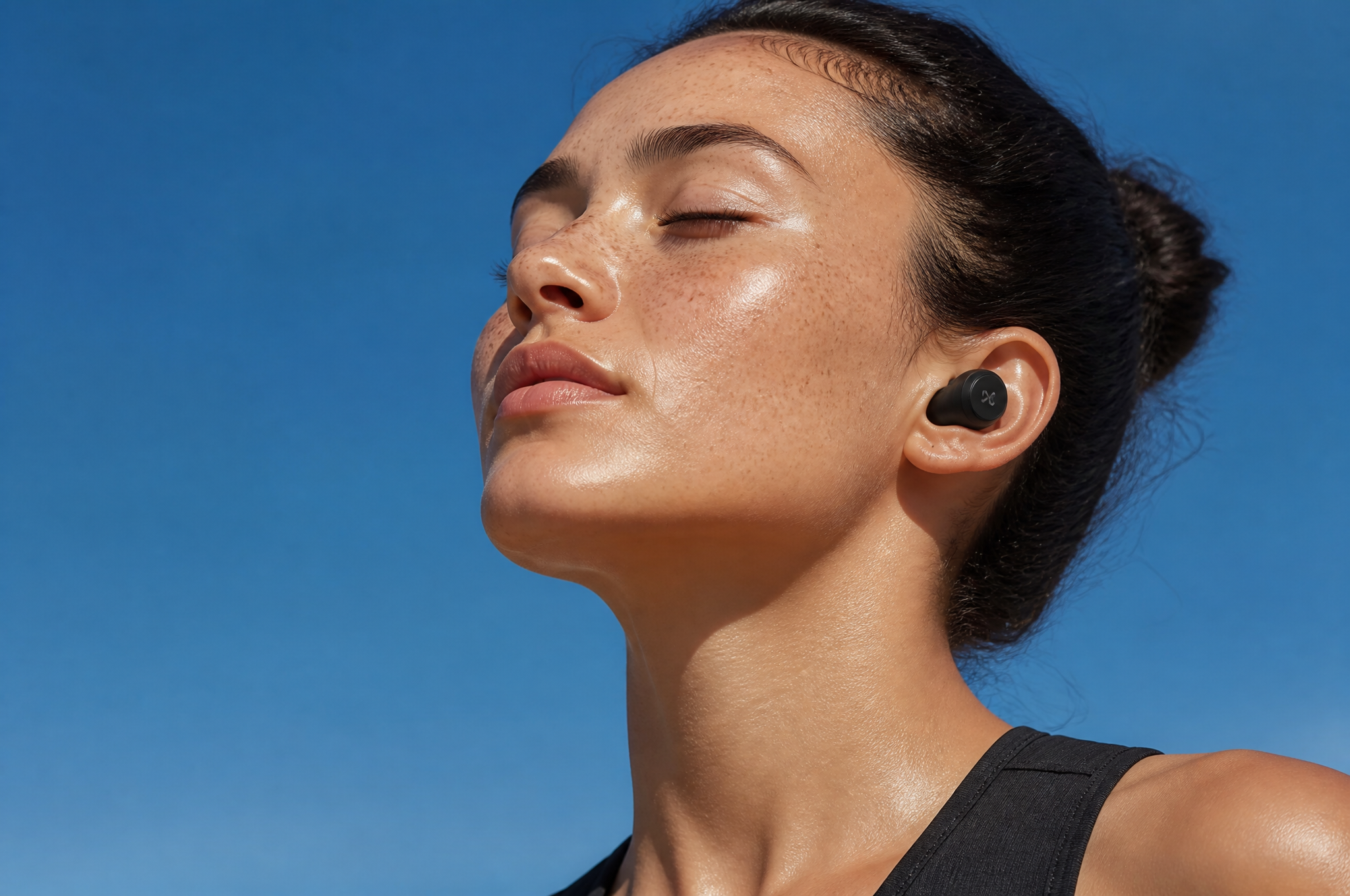A serene close-up of a freckled woman with wireless earbuds, eyes closed, against a clear…