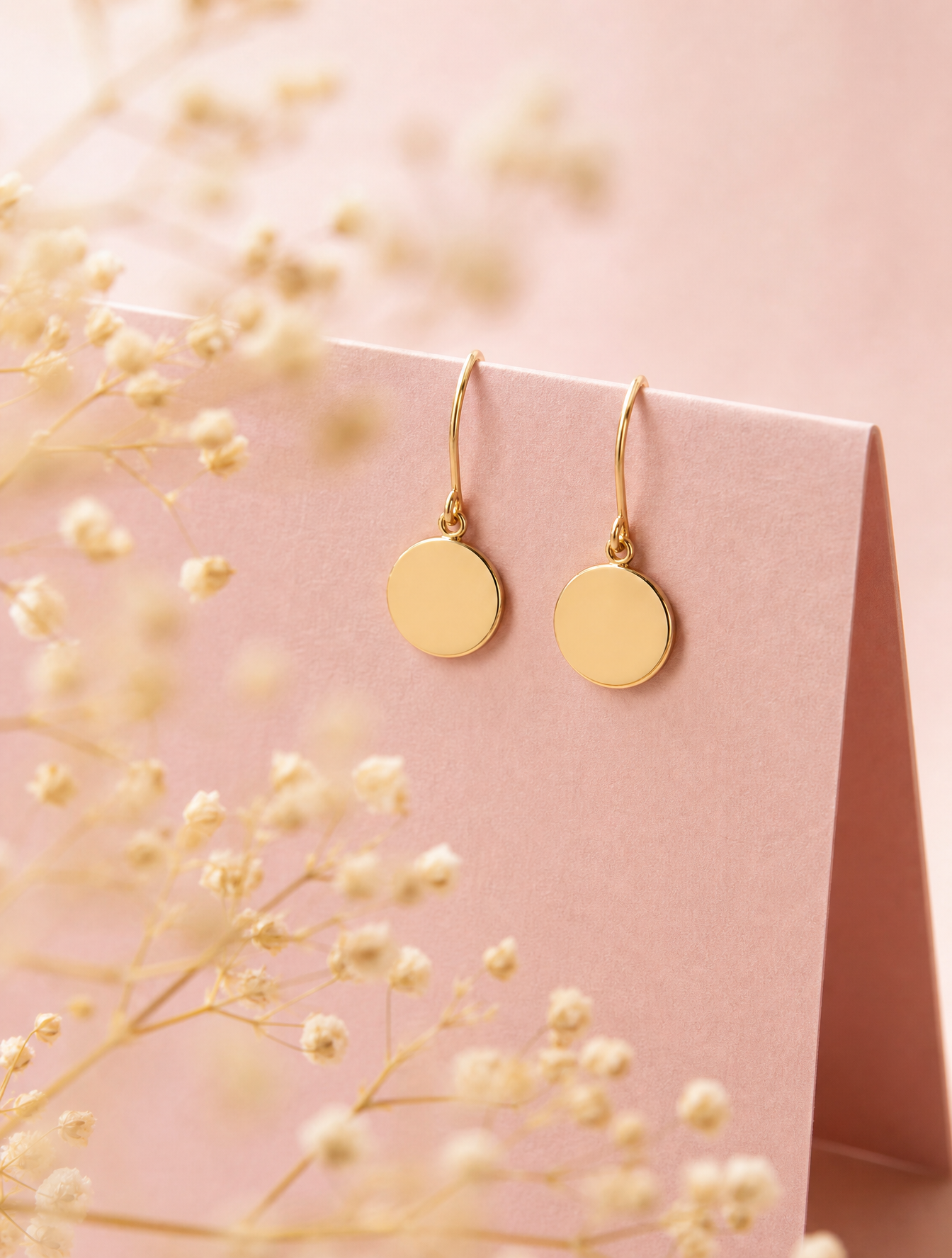 A pair of minimalist gold disc earrings hang delicately from the edge of a folded, matte-…