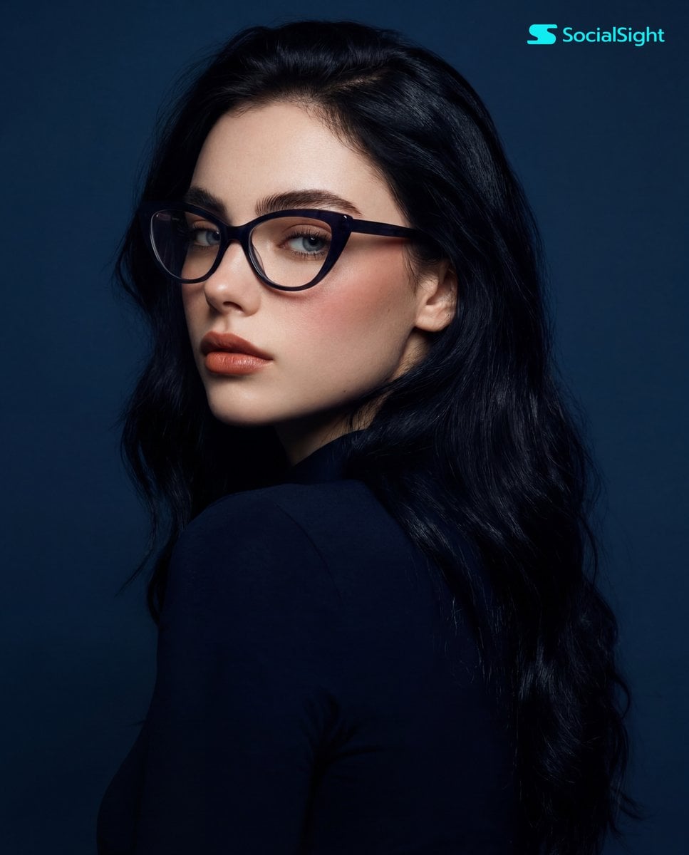 A young woman with long, dark hair and striking cat-eye glasses is captured in a studio p…